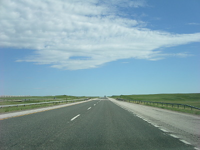 [View taken through the windshield of the vehicle at the roadway ahead. It's flat land with green grass on either side and a huge blue sky overhead with a patch of very white clouds.]